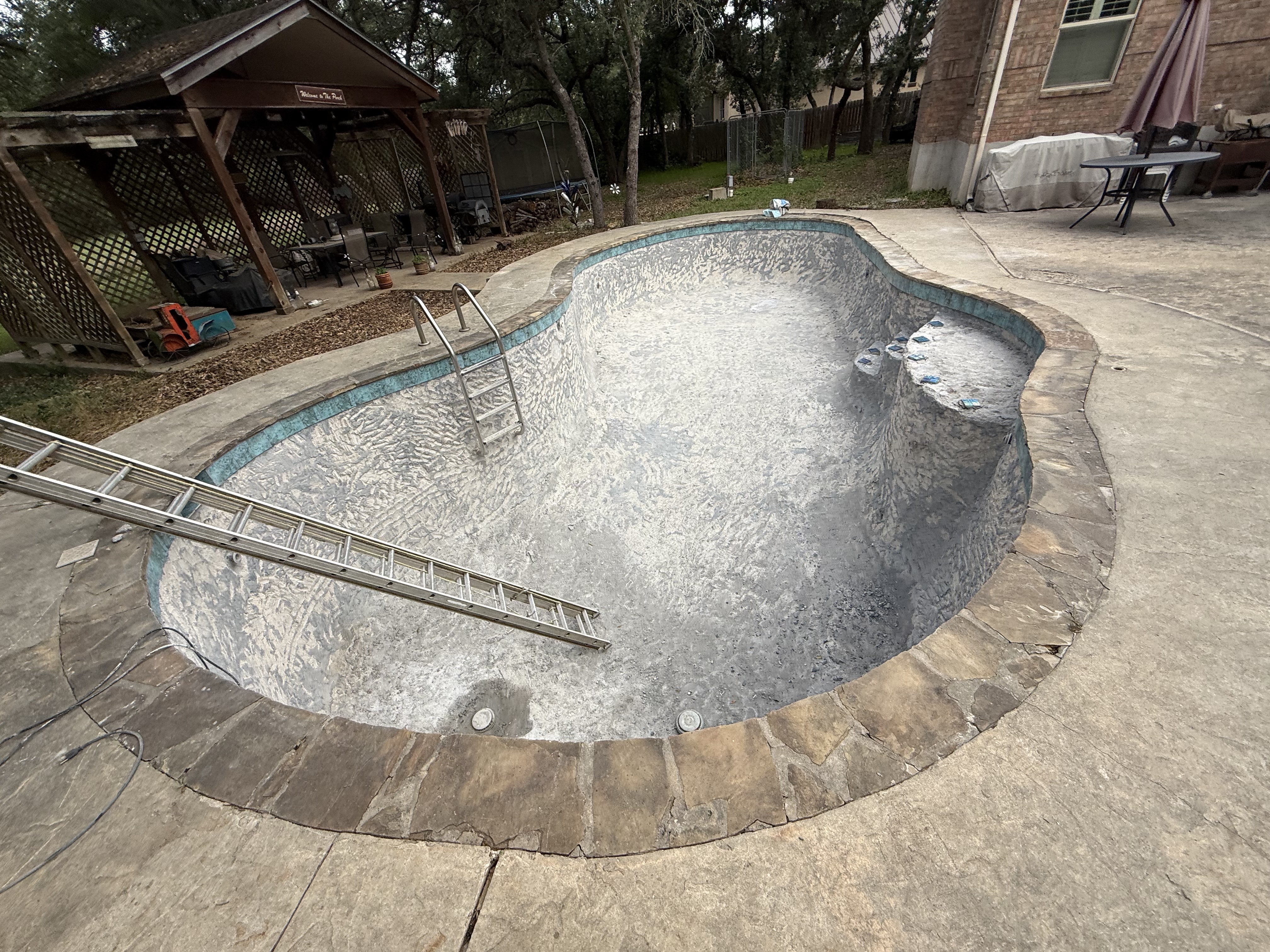Empty pool shell with rough gray plaster chipped away from the walls during the demolition stage of a tile and plaster remodel, with a ladder leaning in and the waterline tile band visible