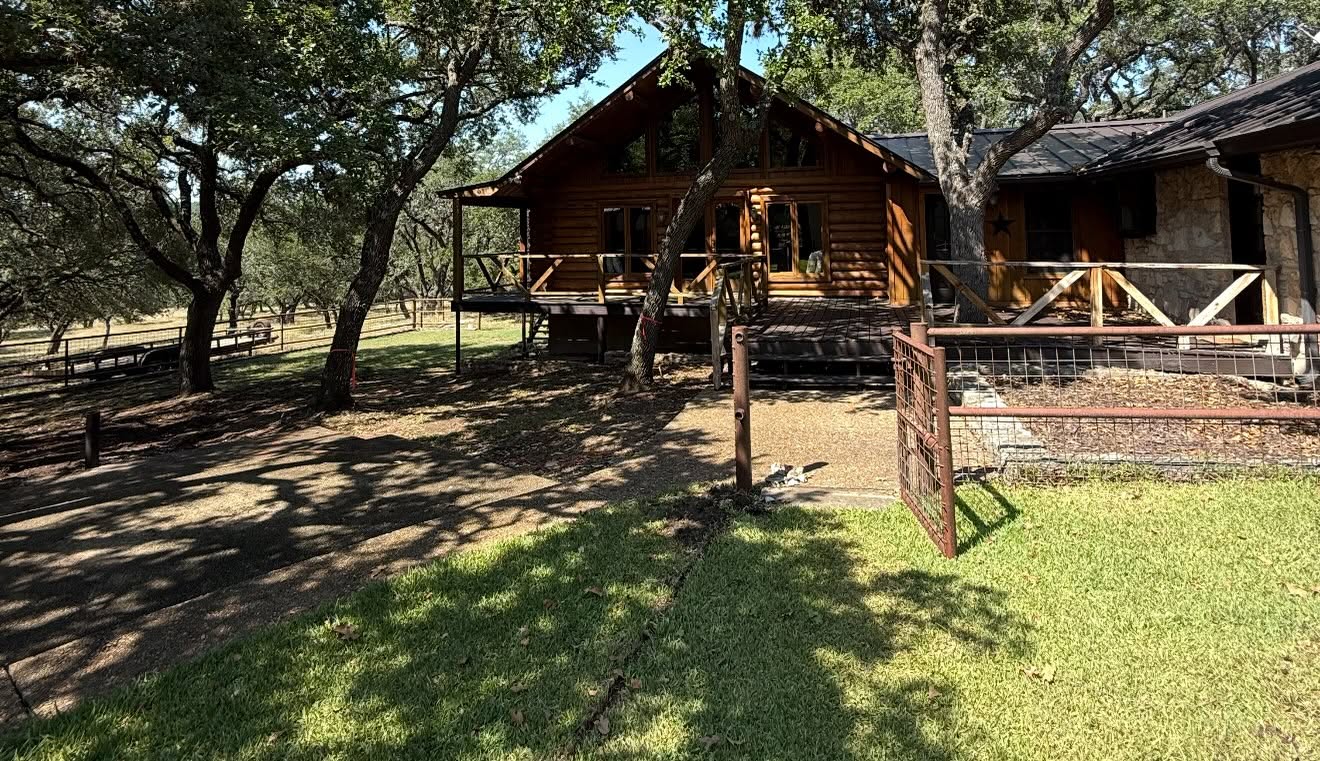 Backyard of a two-story log-cabin home with a weathered wooden deck and mature oak trees before custom pool construction by Asante Pools