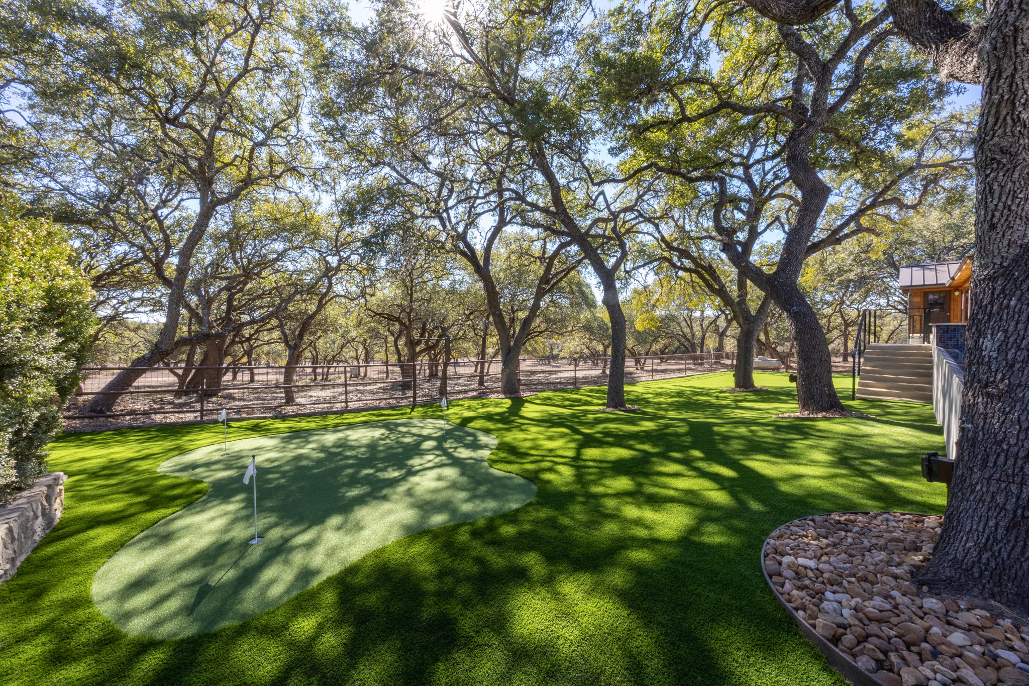 Custom putting green on manicured turf shaded by mature oaks, with stone work around the tree bases and a low retaining wall