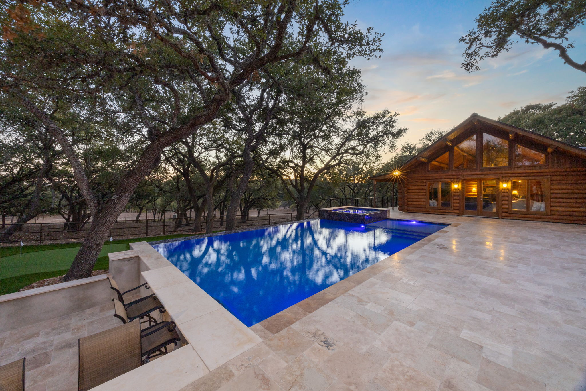 Dusk view of a rectangular infinity pool with raised spa, sunken bar and light stone patio beside a log home, with a small putting green