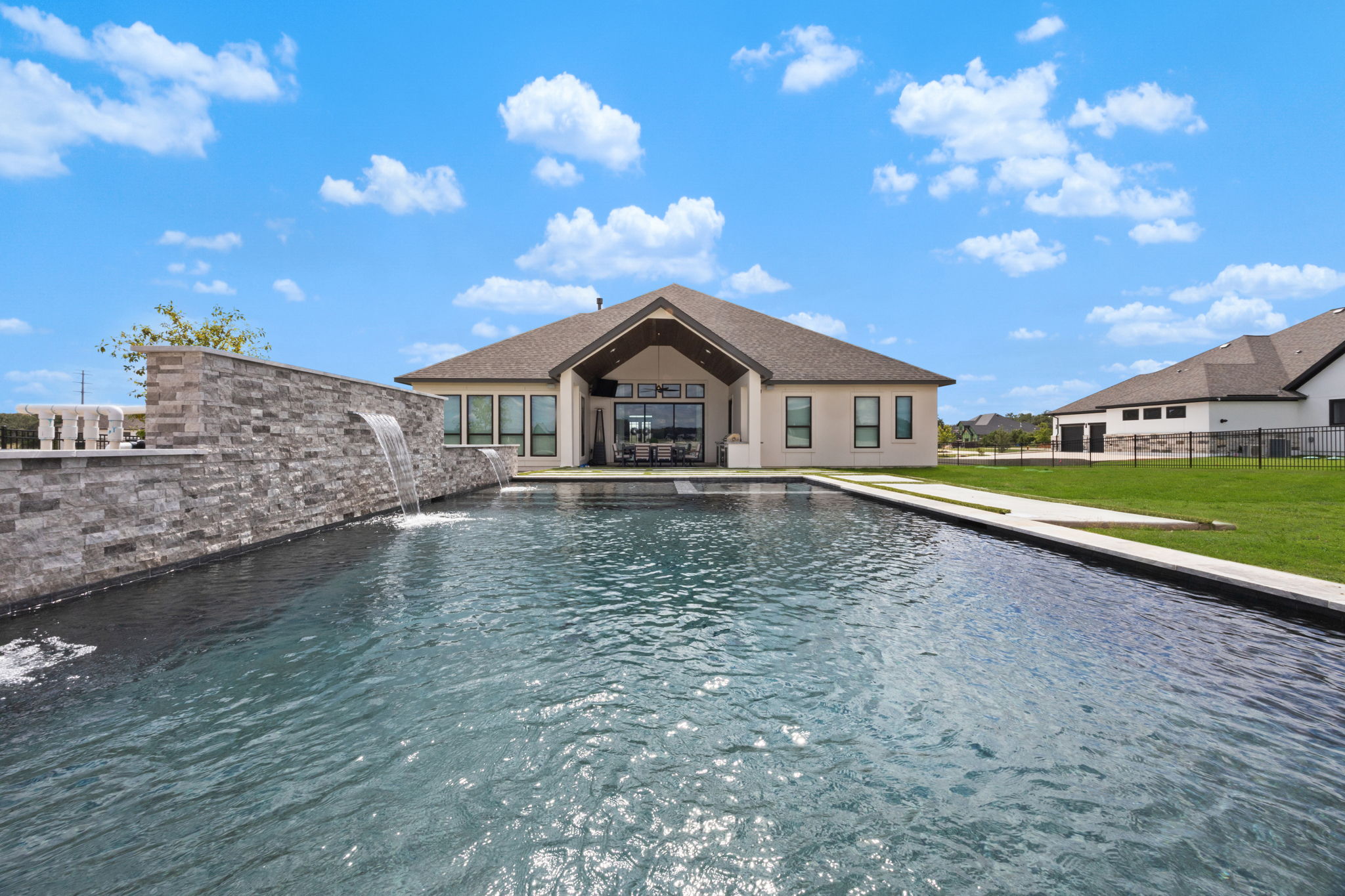 Boerne rectangular dark-bottom pool with two sheer descent waterfalls on a grey stacked-stone wall and paver decking leading to the covered patio