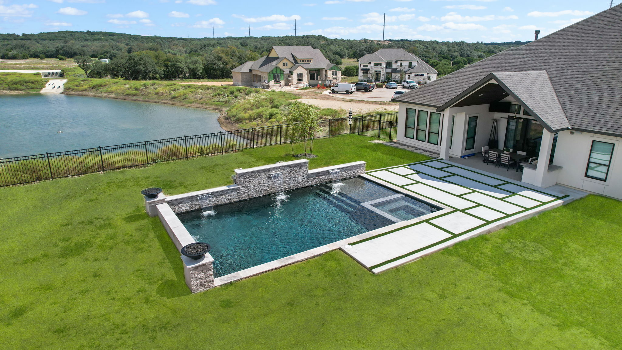 Boerne rectangular dark-bottom pool with an integrated square spa, three sheer descent waterfalls, fire bowls and a concrete paver deck with grass insets overlooking the water