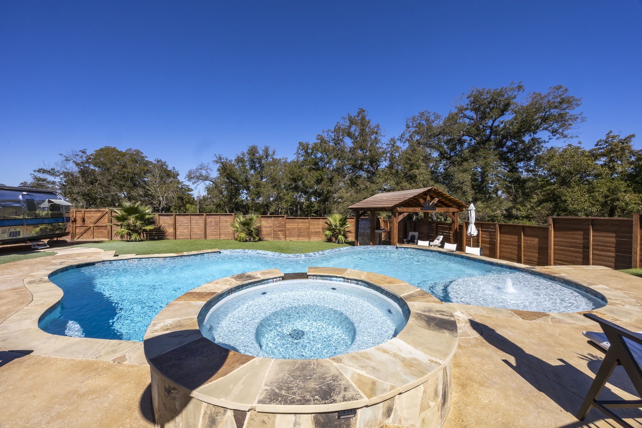 Comfort freeform pool with blue waterline tile, a light finish, a tanning ledge bubbler and an integrated raised circular stone spa framed by a wooden pavilion and mature trees