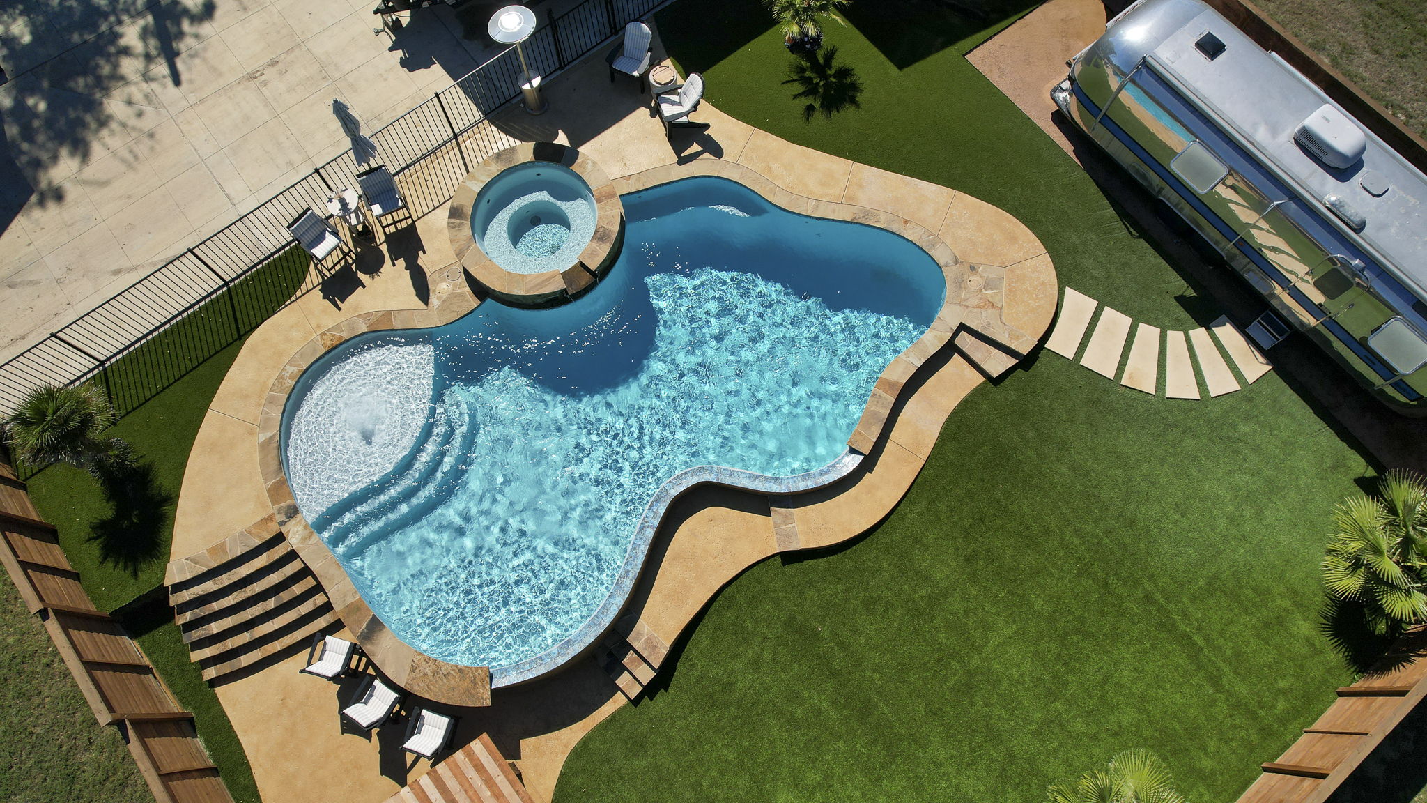 Comfort aerial view of a freeform light blue pool with a tanning ledge bubbler, raised circular spa, blue waterline tile and tan stone coping set in artificial turf