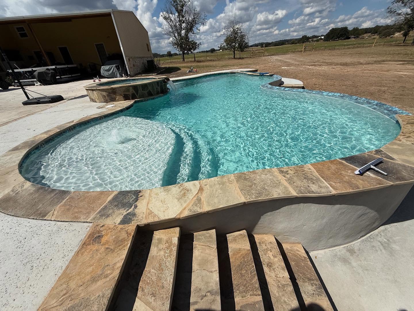 Freeform pool with a flagstone-coped tanning ledge and bubbling fountain beside a raised stacked-stone spa with spillover waterfall on a brushed concrete deck in a rural Comfort backyard