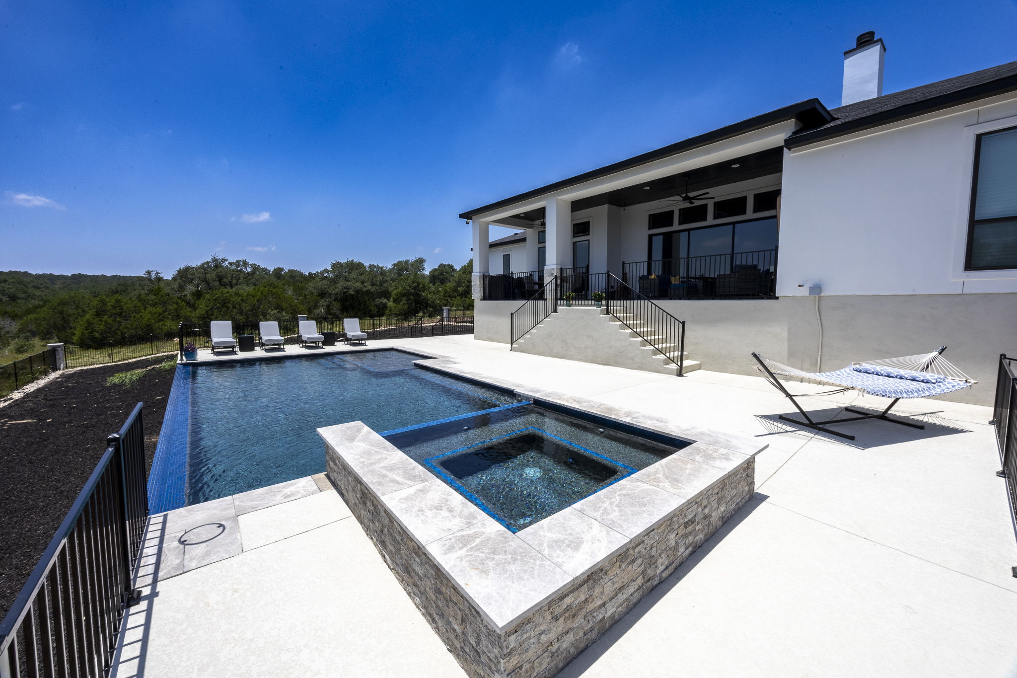 New Braunfels pool with a dark tiled interior, integrated square spa and lounge chairs overlooking rolling Hill Country views