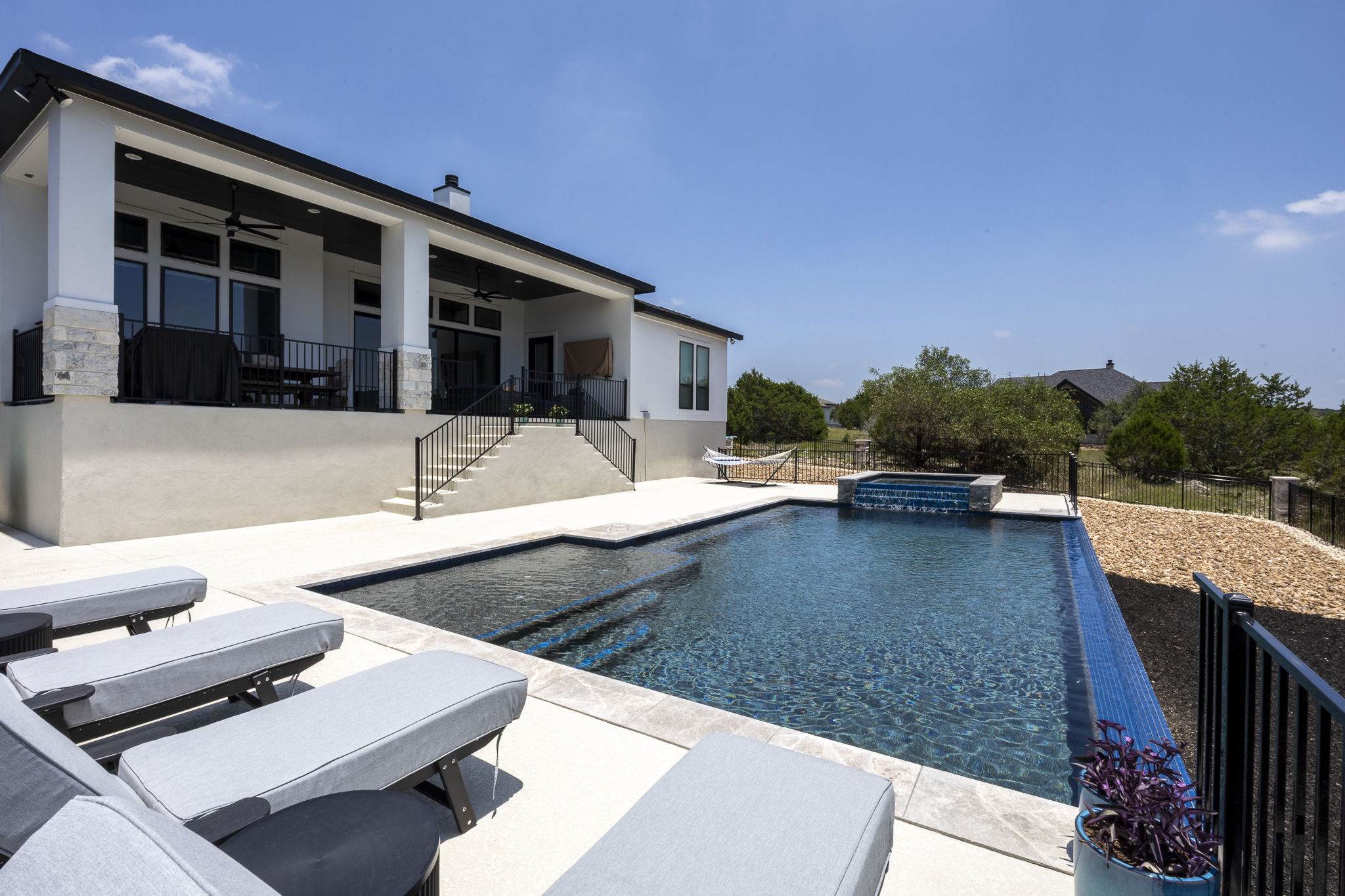 New Braunfels rectangular pool with a sun shelf, raised stone spa waterfall spillover and lounge chairs beside the covered patio