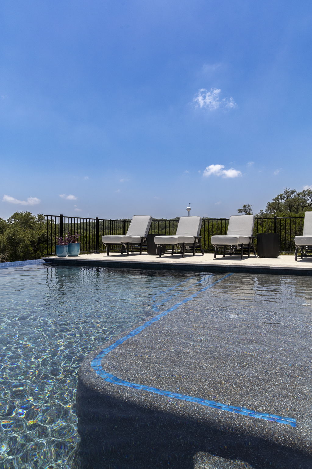 New Braunfels pool with a pebble finish, blue-tiled sun ledge and white lounge chairs along a stone deck