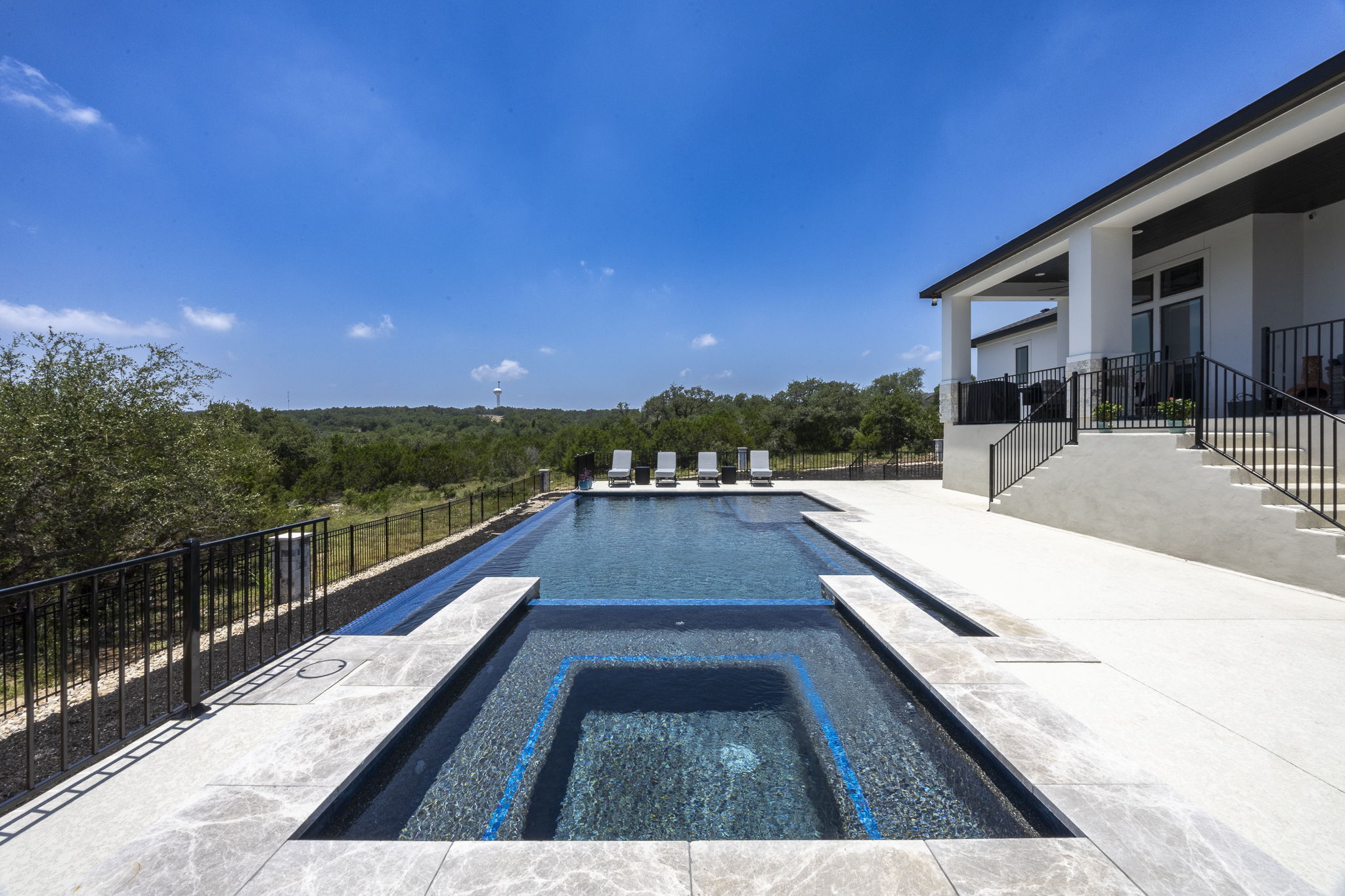 New Braunfels square spa with blue tile accents beside a long rectangular infinity-edge pool below the covered porch