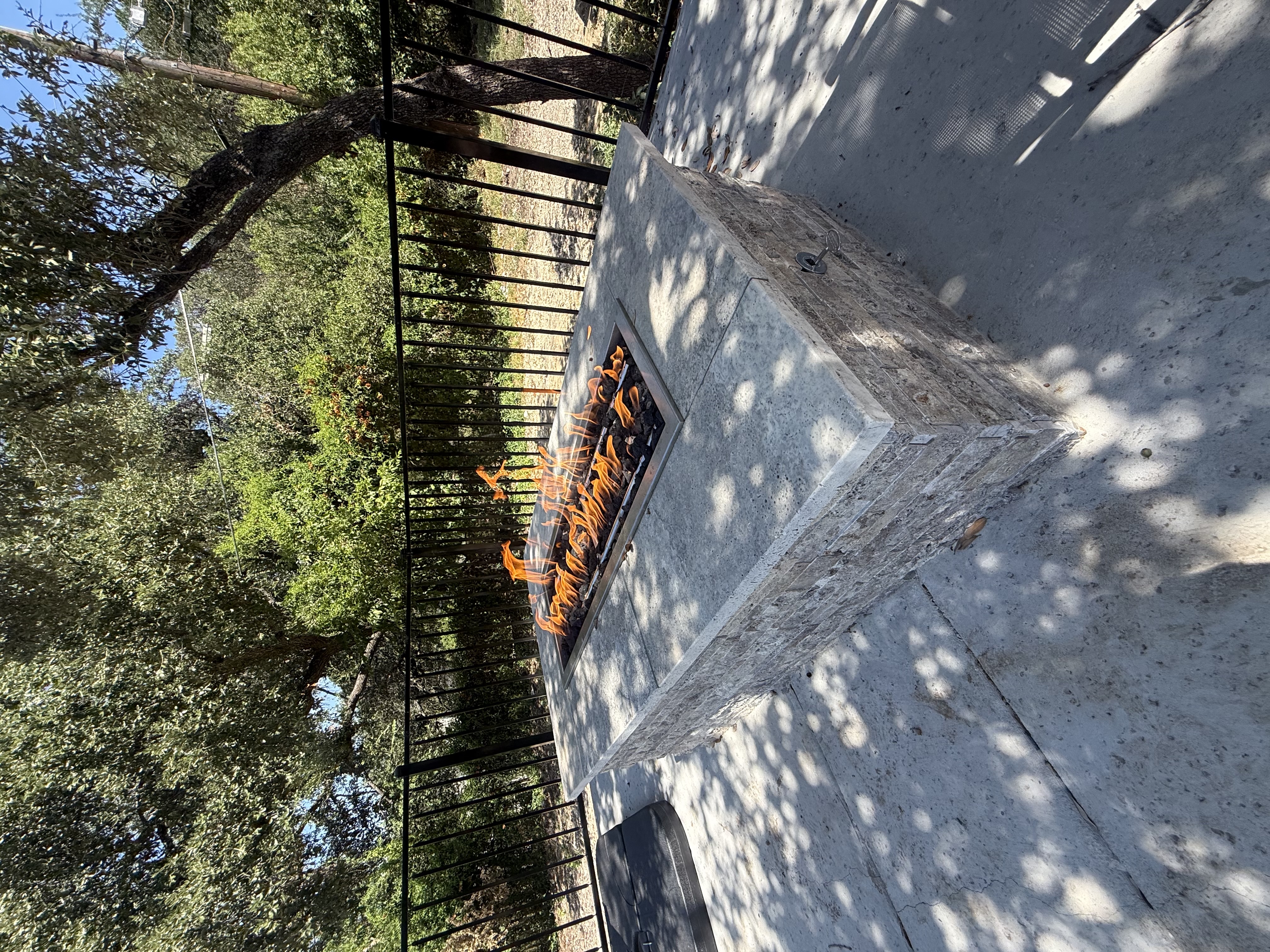 Rectangular stone fire pit with active flames and a smooth slab top on a concrete patio bordered by a black metal fence